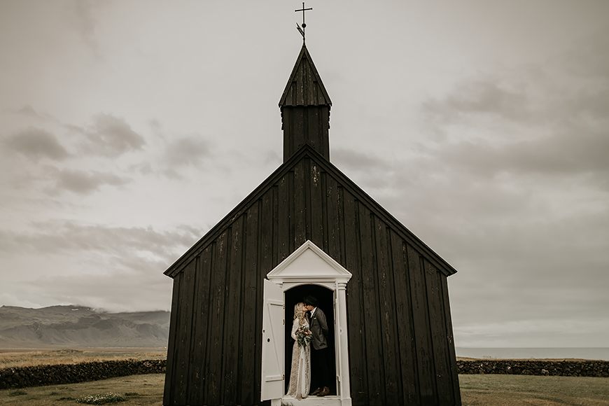 A bride and groom standing in front of a black church in iceland.