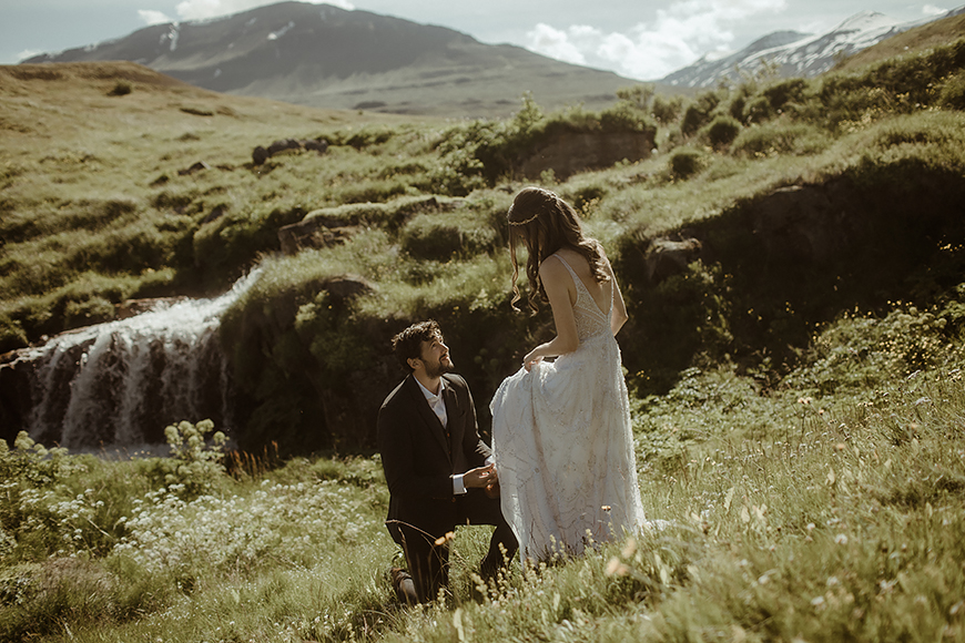 A bride and groom standing next to a waterfall in the mountains.