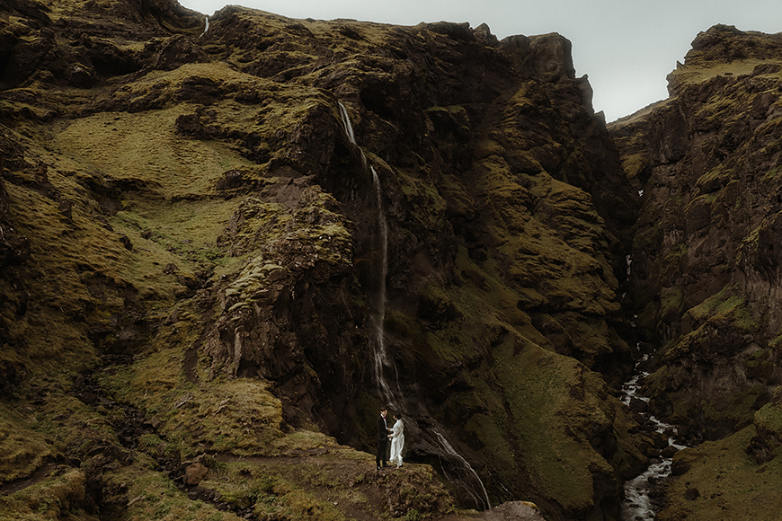 A bride and groom standing on a cliff overlooking a waterfall.