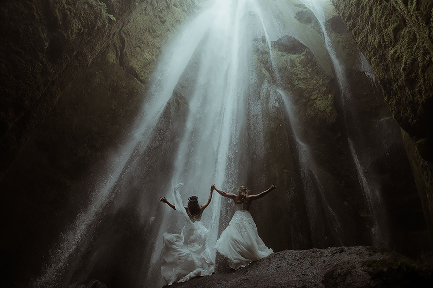 Two brides standing in front of a waterfall.