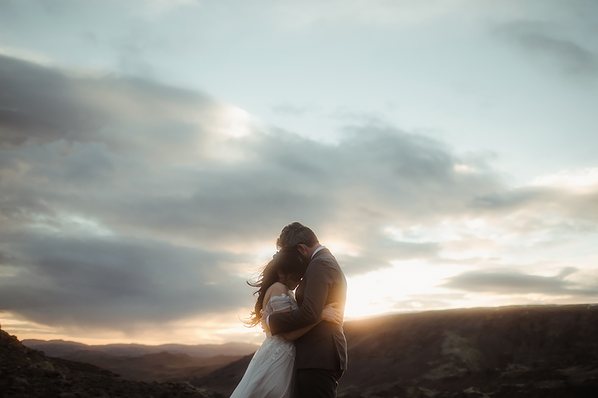 A bride and groom embrace on top of a mountain at sunset.