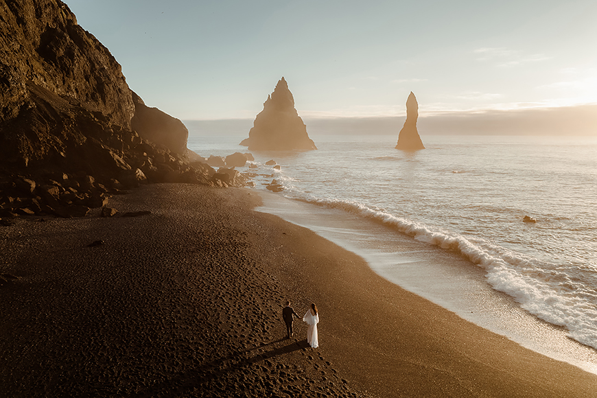 A bride and groom standing on a beach with cliffs in the background.