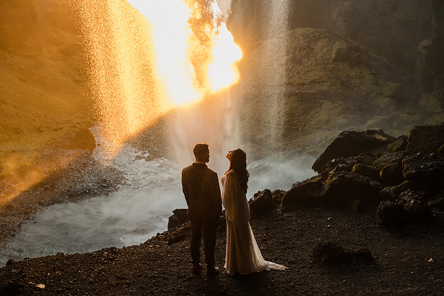 A bride and groom standing in front of a waterfall at sunset.