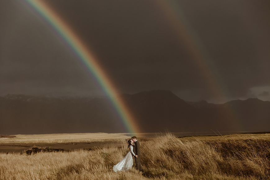 A bride and groom standing under a rainbow in a field.